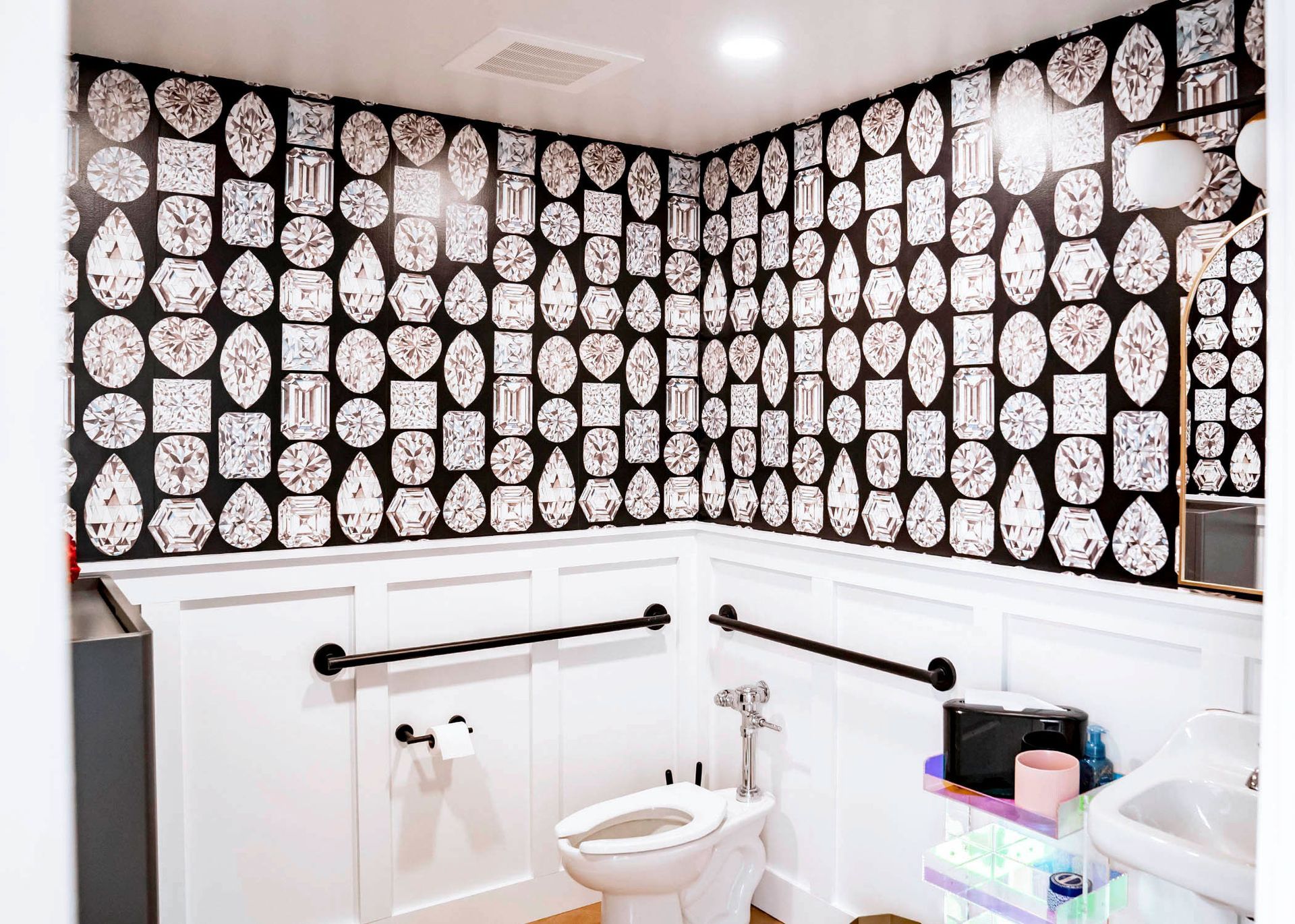 Bathroom with diamond-patterned wallpaper, white wainscoting, and grab bars. Toilet is in view.