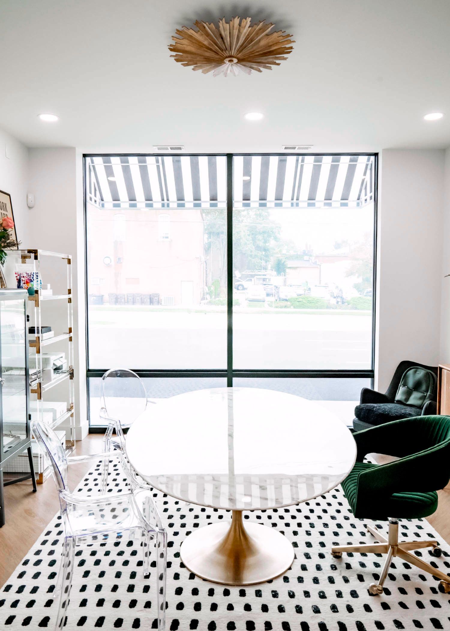 White office with round table, black and white rug, green chair, striped awning, and large window.