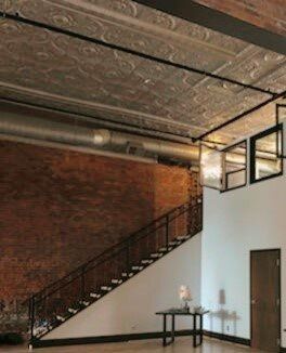 Industrial-style interior with exposed brick wall, metal staircase, tin ceiling, and open ductwork.