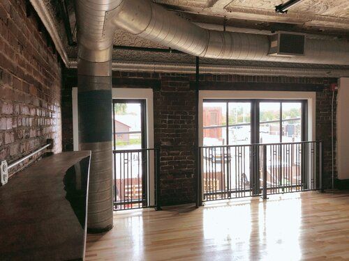 Interior of a brick-walled loft space with large windows, balcony, and exposed ductwork.