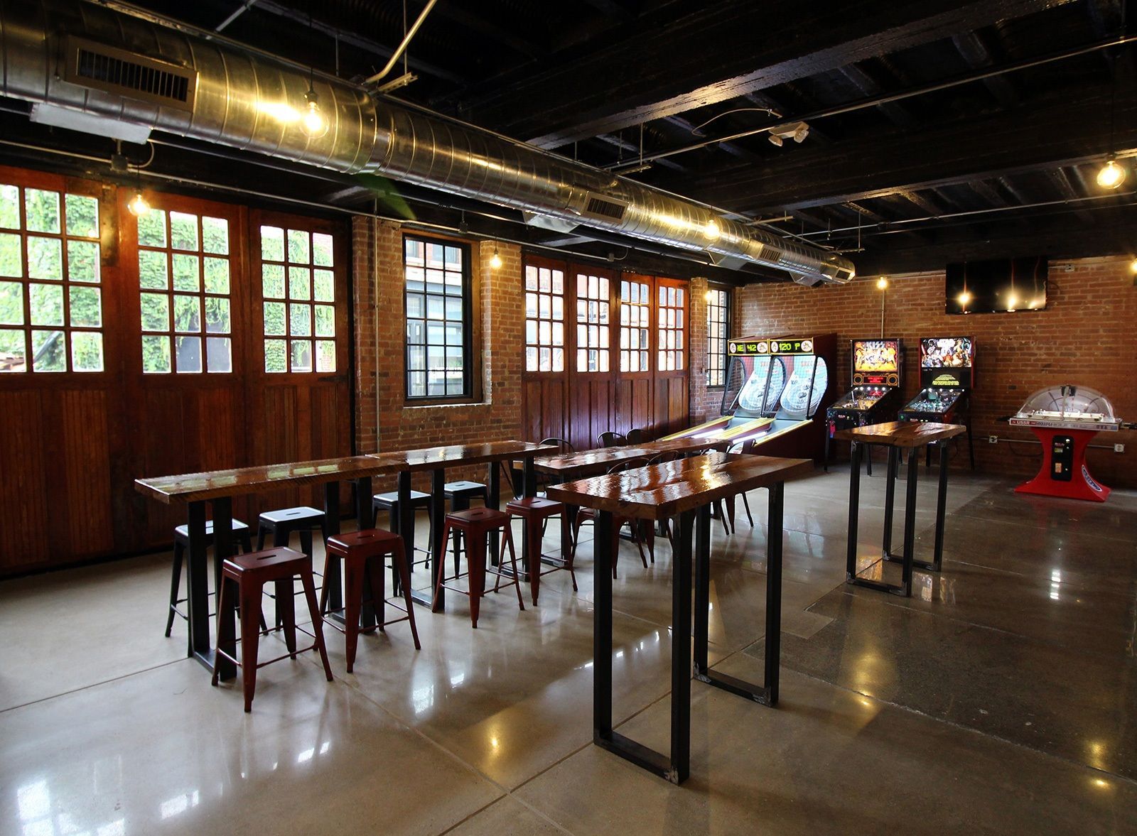 Interior of a room with brick walls, arcade games, long tables, and stools. Large wooden doors and windows are along the back wall.