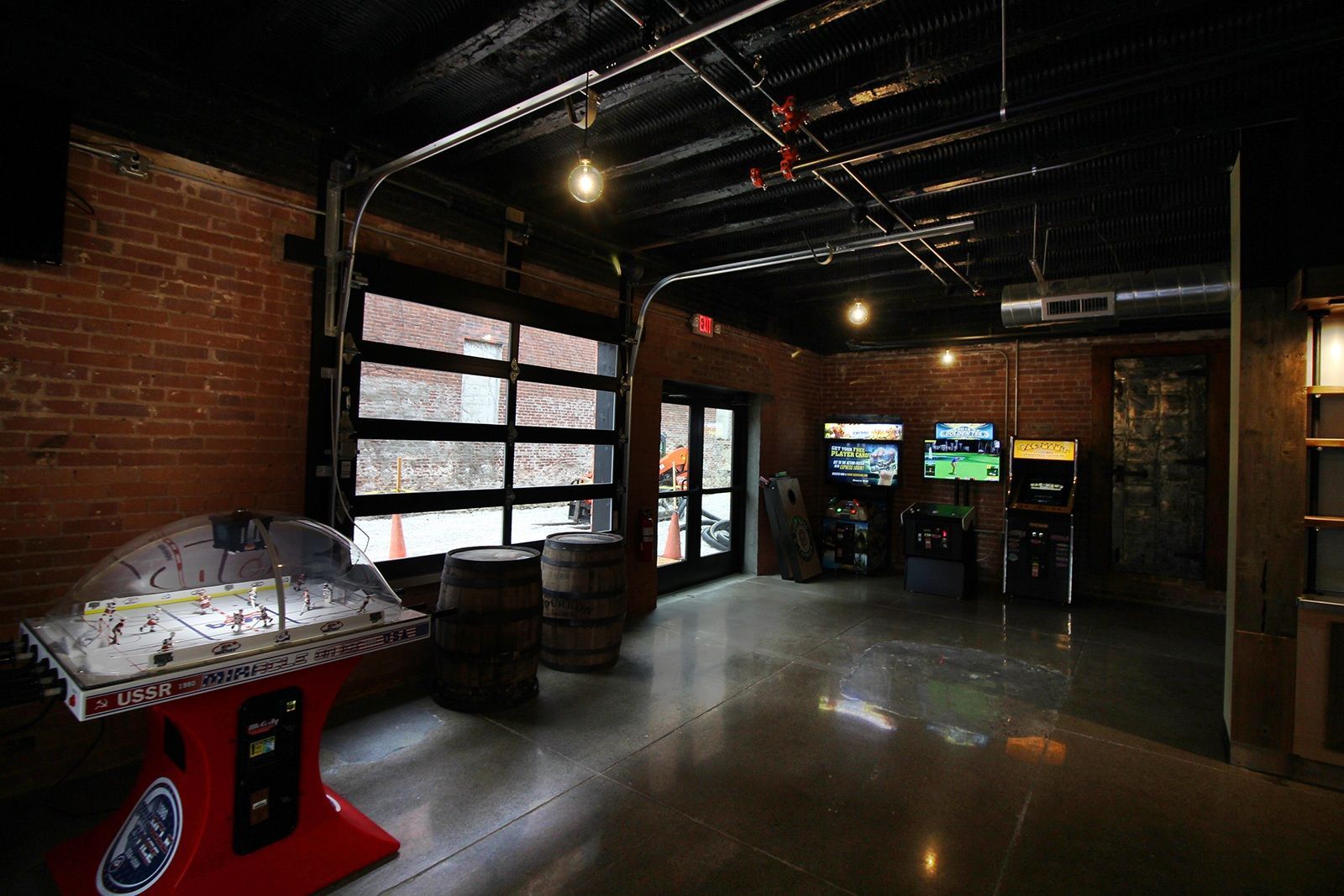Interior of an arcade with brick walls, exposed pipes, garage door, arcade games, and air hockey.