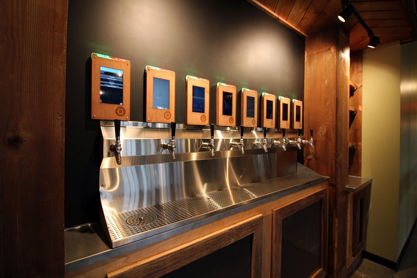 Beer tap system with digital screens, stainless steel spouts, and wooden accents in a bar setting.