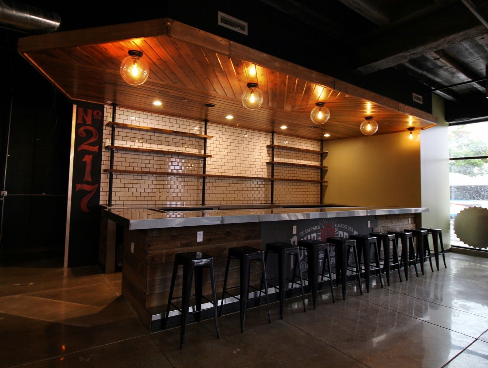 Bar with black stools, wooden countertop, shelves against a white brick wall, and globe lights.