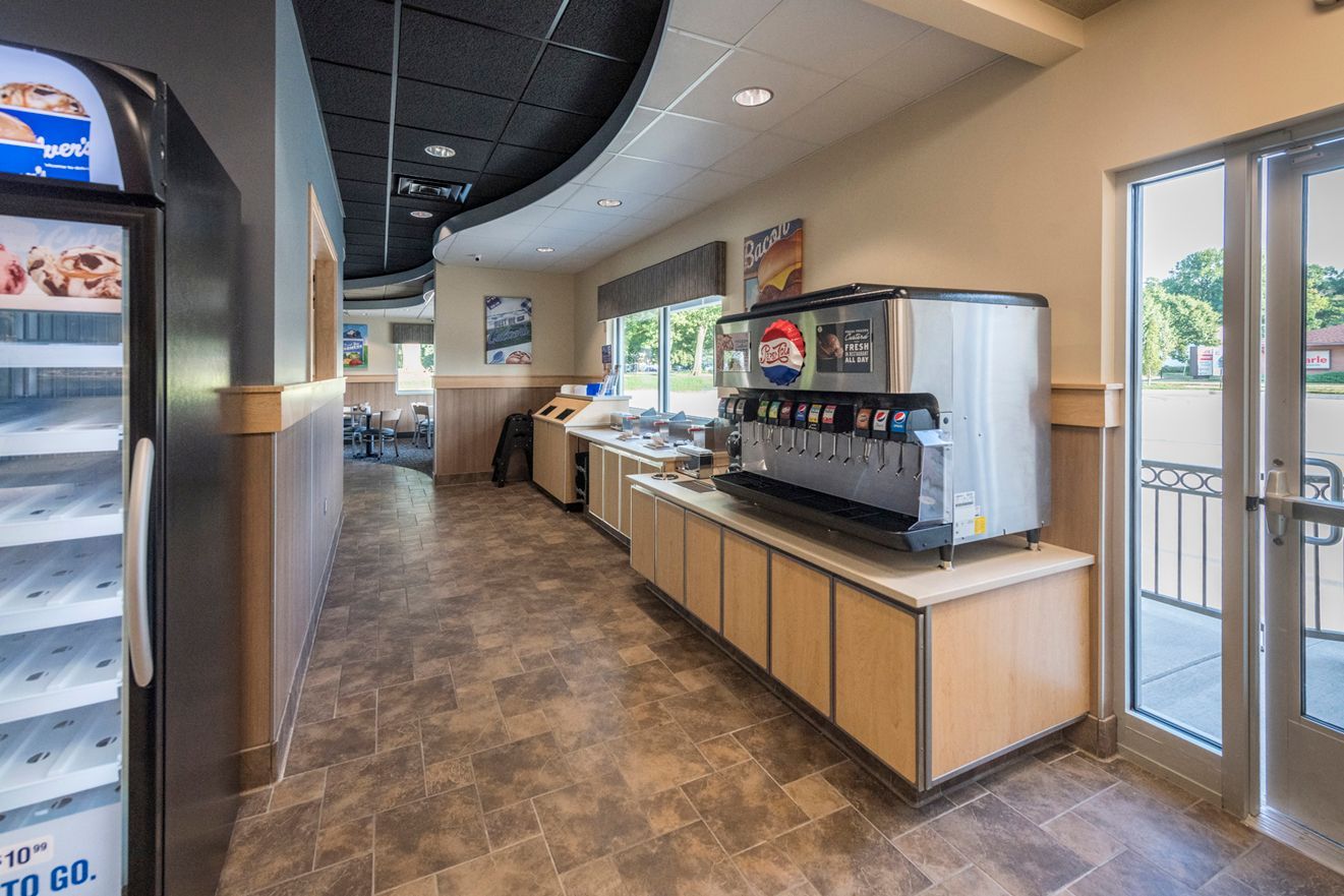 Interior of a fast-food restaurant with a drink dispenser, cooler, and service area. Beige and brown tones are dominant.