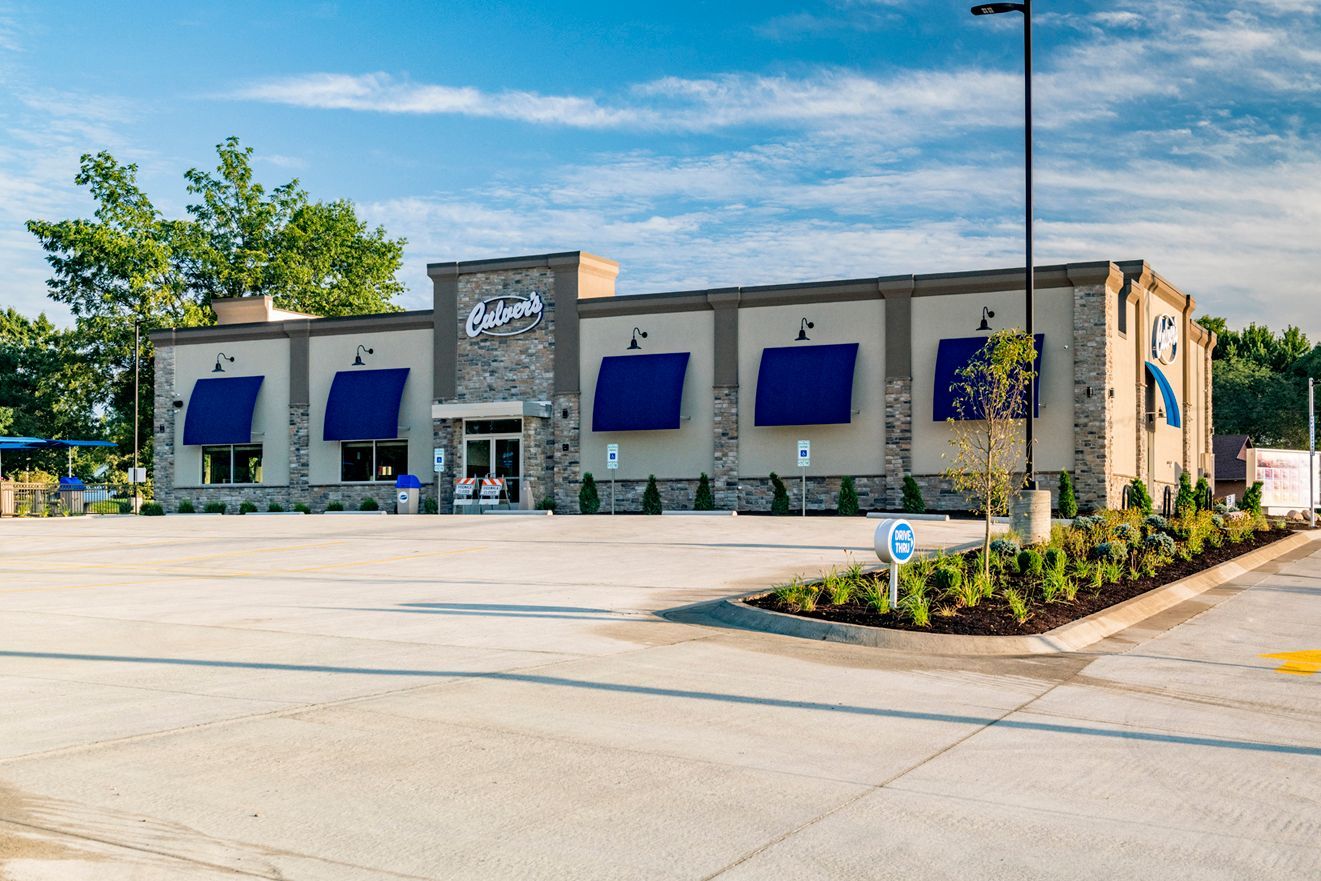 Exterior of a Culver's restaurant, tan building with blue awnings. Empty parking lot, sunny day.