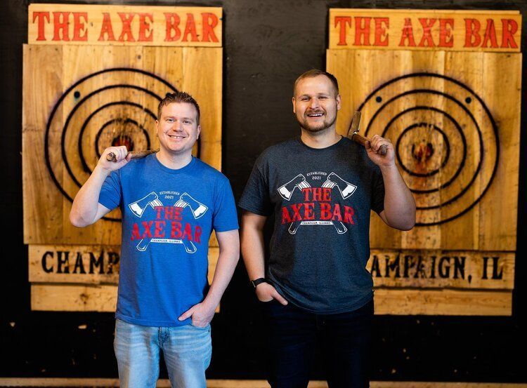 Two men holding axes in front of targets at The Axe Bar in Champaign, IL. Both wear Axe Bar shirts.
