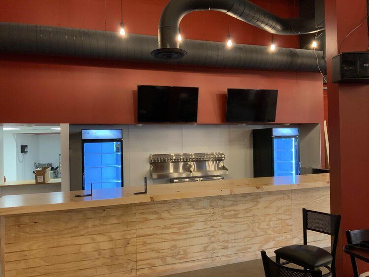 Bar interior: wooden bar with beer taps, two TVs on red wall, two blue-lit refrigerators, and ventilation.