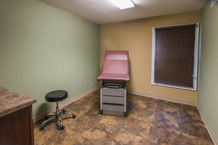 Medical examination room with exam table, stool, and small window with blinds.