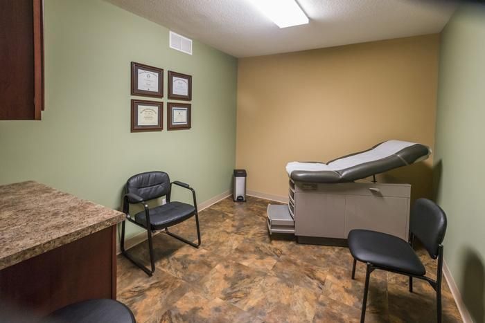 Medical exam room with examination table, chairs, and framed artwork on green and tan walls.