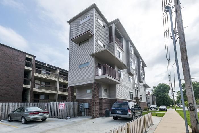 Modern multi-story building with balconies, gray siding, and parked cars on a driveway.