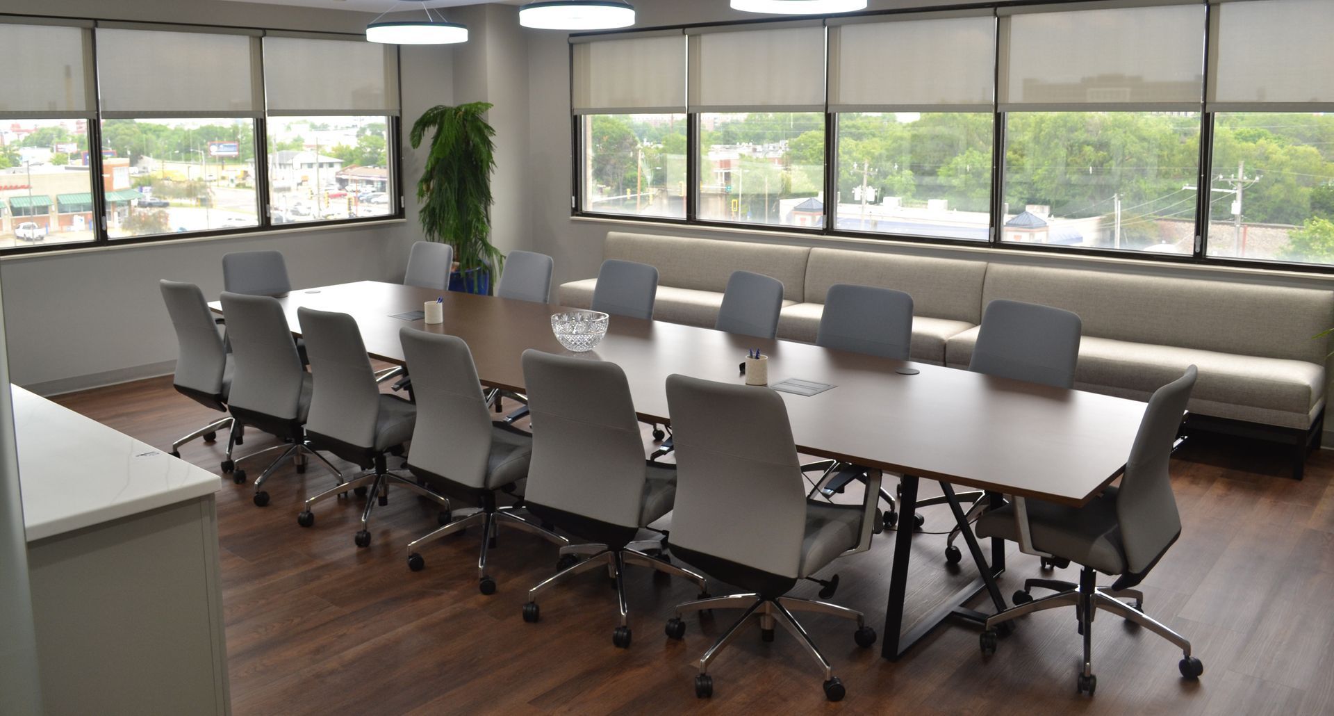 Empty modern conference room with long table, gray chairs, large windows, and wood floor.