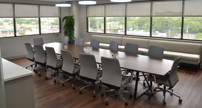 Empty modern conference room with long table, gray chairs, large windows, and wood floor.