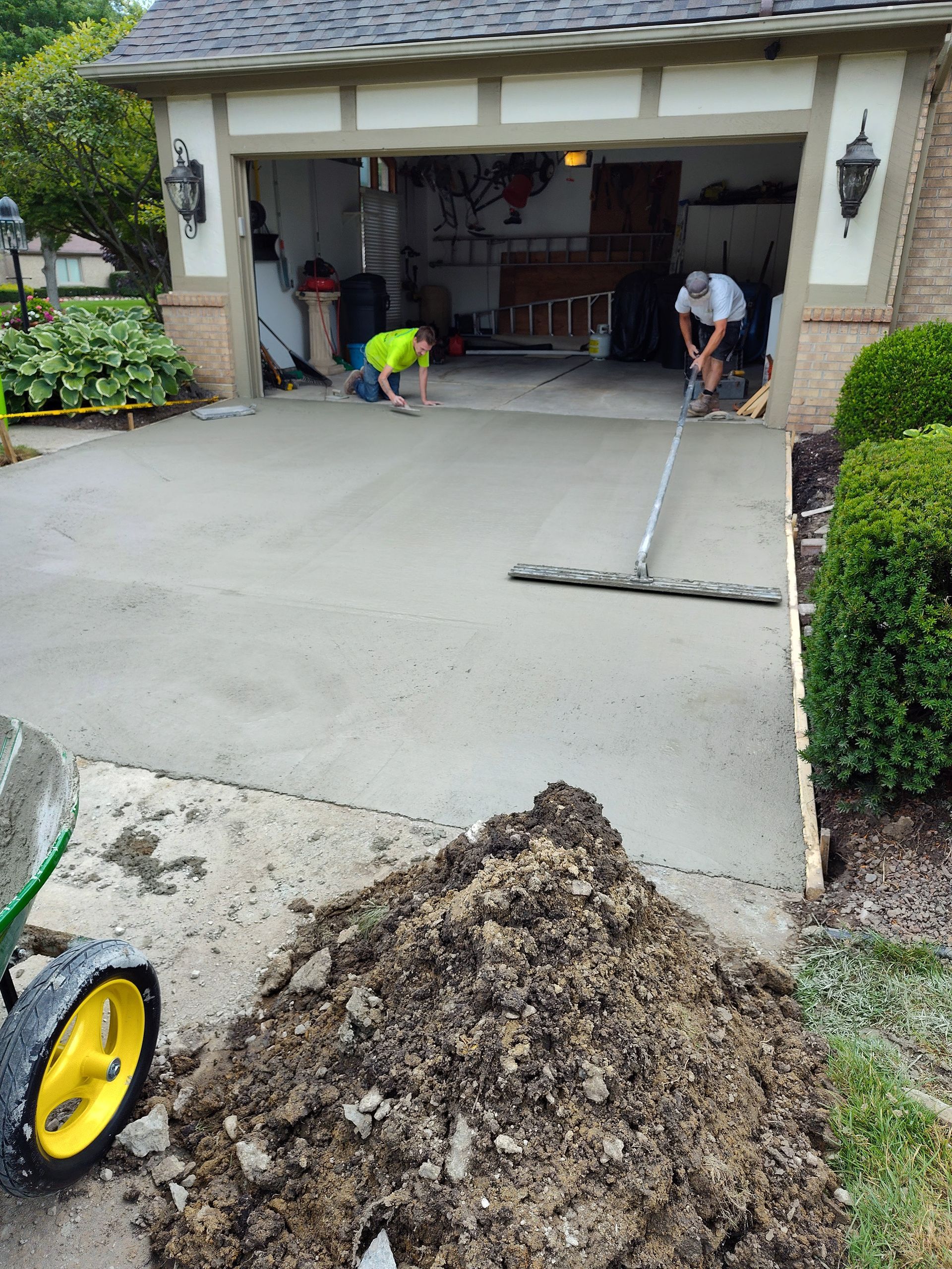 Concrete workers pouring and smoothing a new driveway in front of a garage. A pile of dirt is in the foreground.