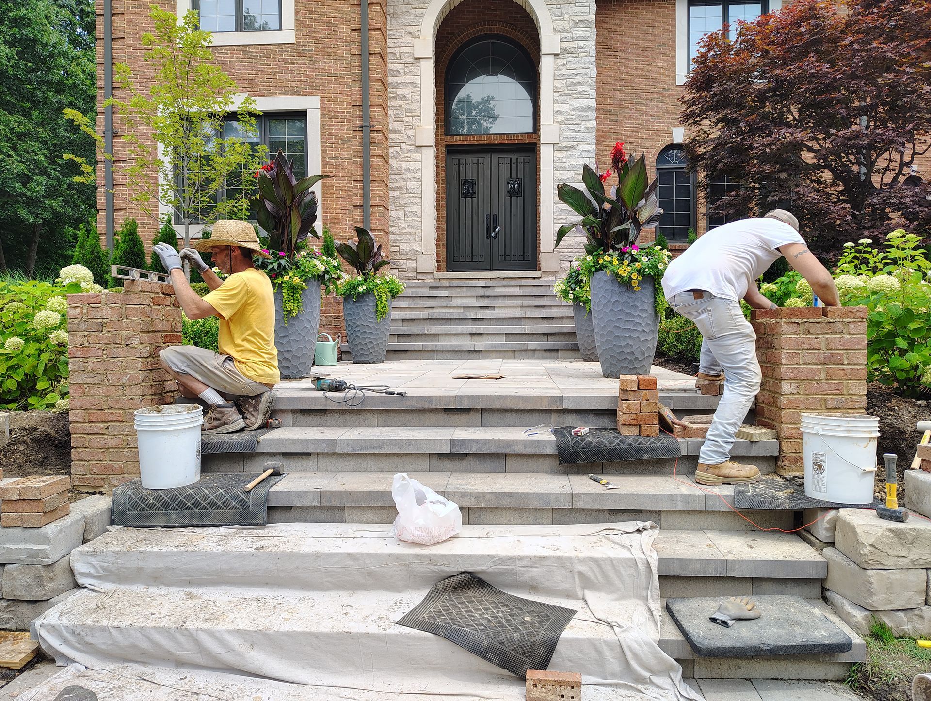 Two workers building brick pillars beside a home's stone steps.