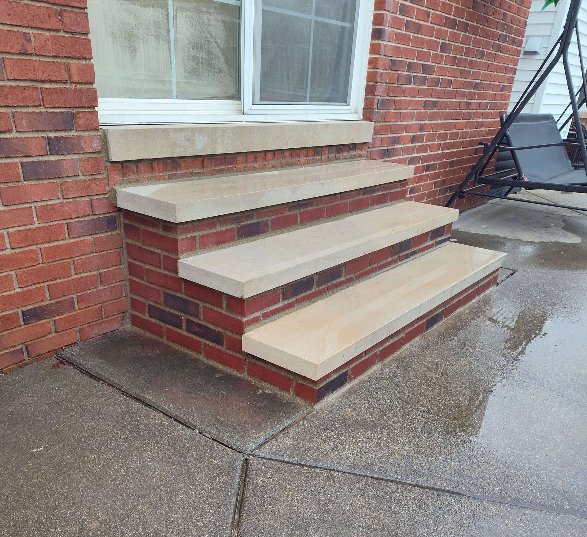 Brick steps with tan stone treads leading to a window. Concrete patio in foreground.