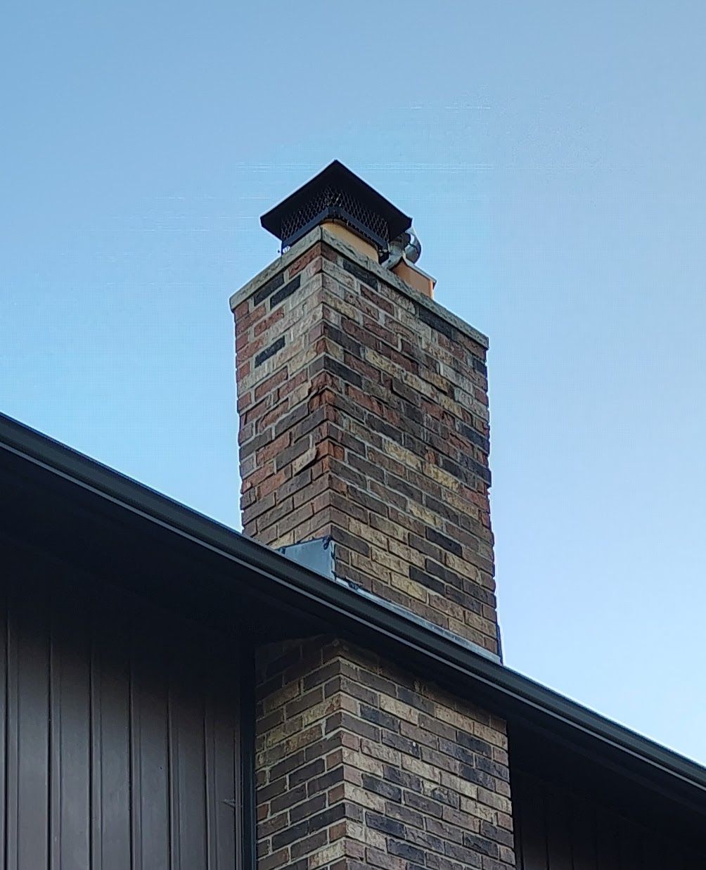 Brick chimney with black cap against a blue sky, rising from a brown roof.