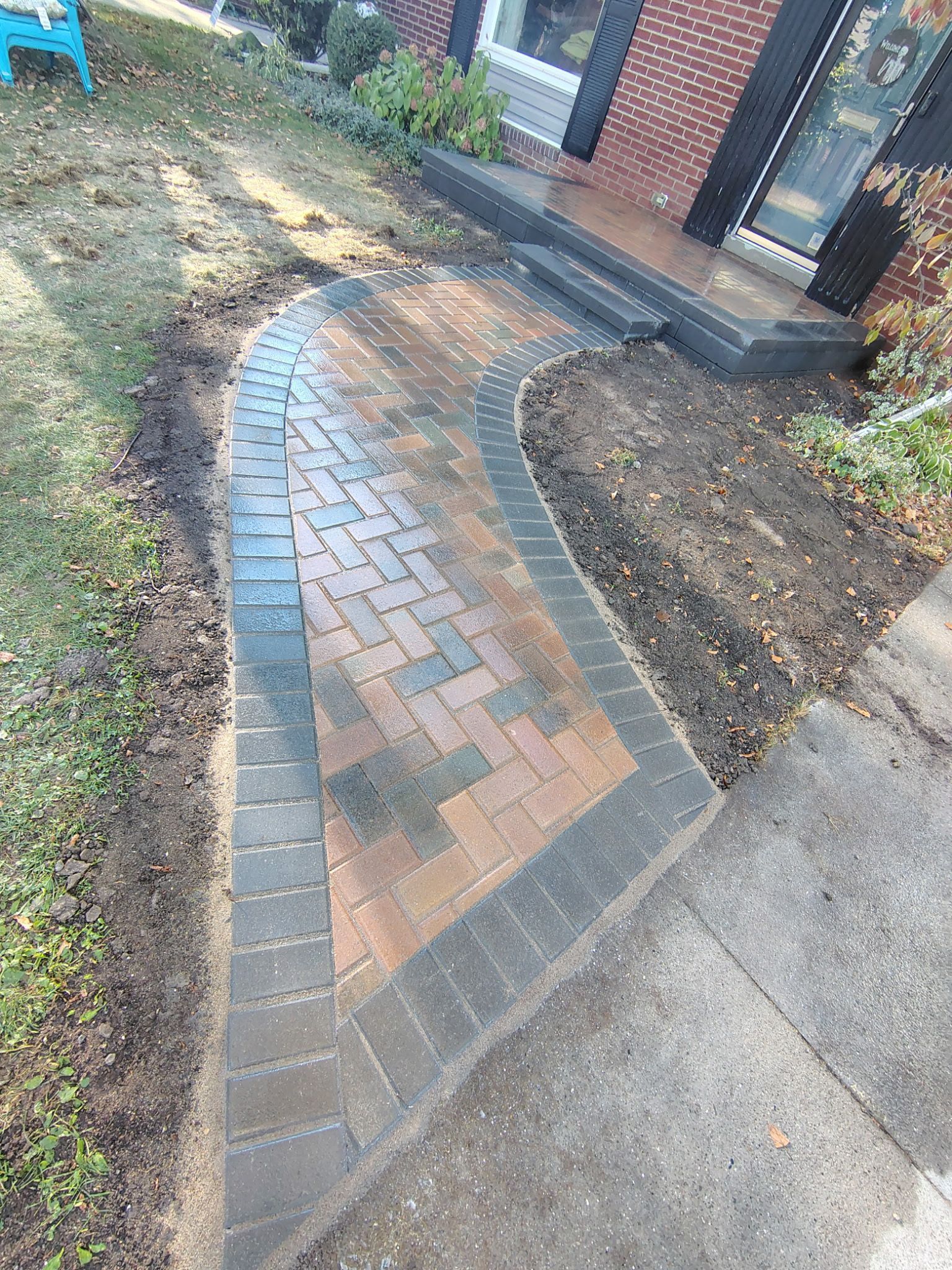 A brick walkway with a dark border leads from a sidewalk to the front steps of a brick house.