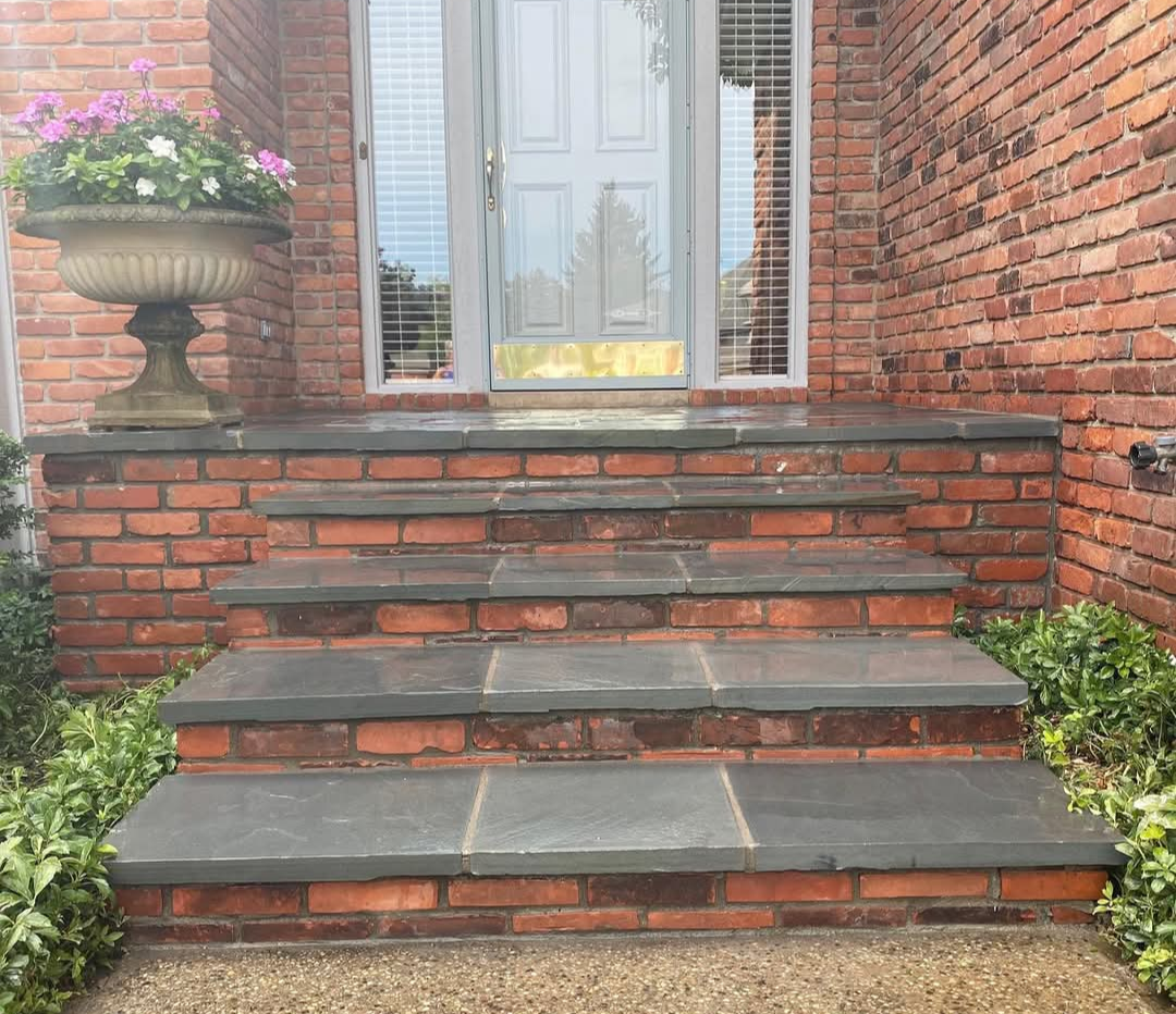 A set of brick steps with dark stone treads leading up to a front door, flanked by a planter with flowers and greenery.