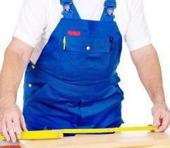 A man in blue overalls is measuring a piece of wood with a tape measure.