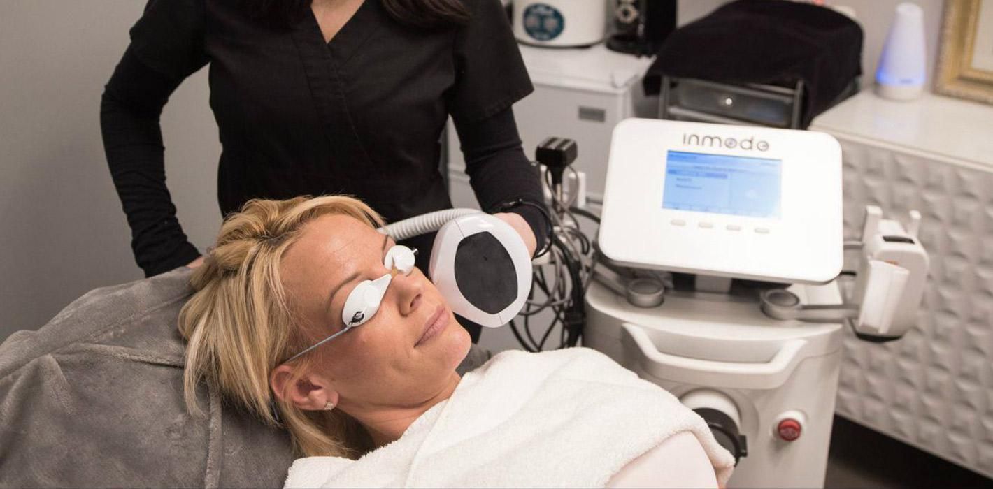 Woman receiving face treatment at a clinic. A technician holds a device near her face.