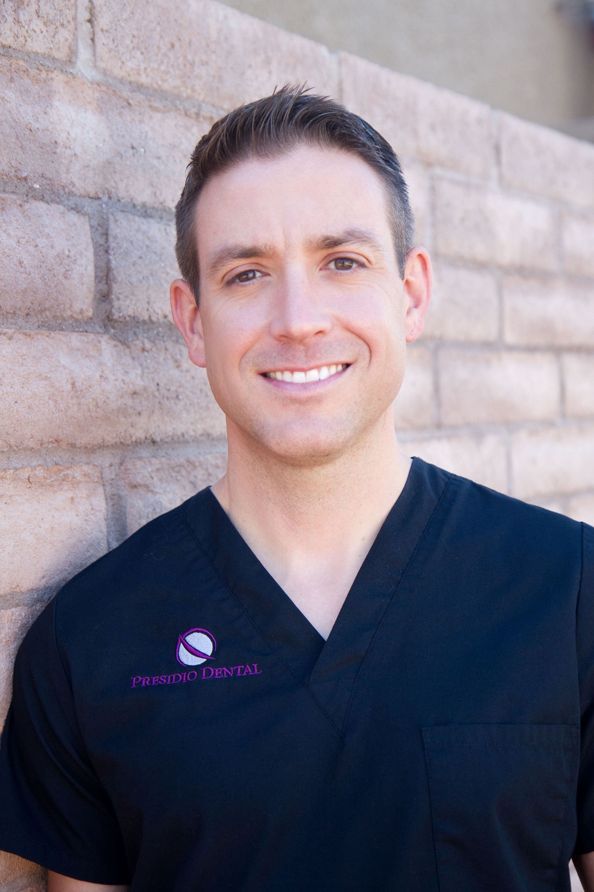 A man in a black scrub top is smiling in front of a brick wall.