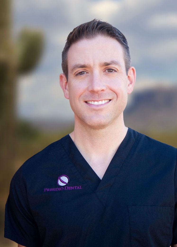 A man in a black scrub top is smiling in front of a brick wall.
