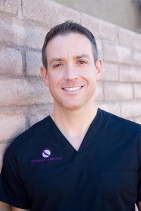 A man in a black scrub top is smiling in front of a brick wall.