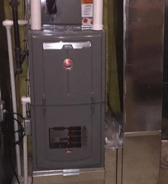 Gray furnace in a utility room with exposed pipes and ductwork.