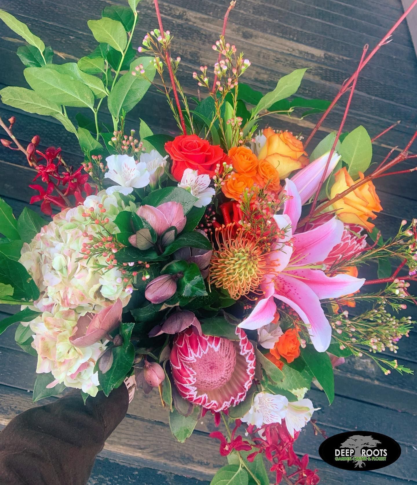 A vibrant floral bouquet with pink lily and protea, orange and red roses, greenery, held against wood.