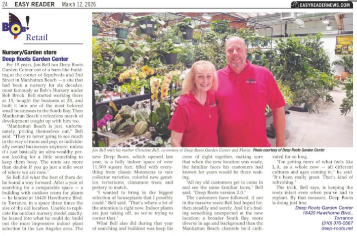 Two people stand smiling inside Deep Roots Garden Center, surrounded by lush potted plants and a greenhouse-style interior.