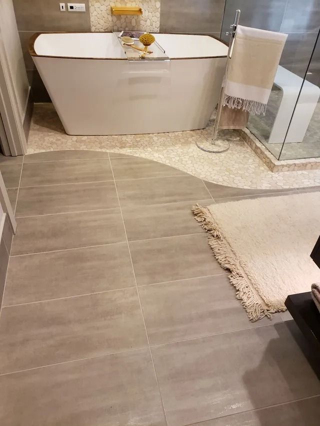 Bathroom with a white freestanding tub and neutral-toned tiled floor; a fluffy rug sits on the floor near the right side.