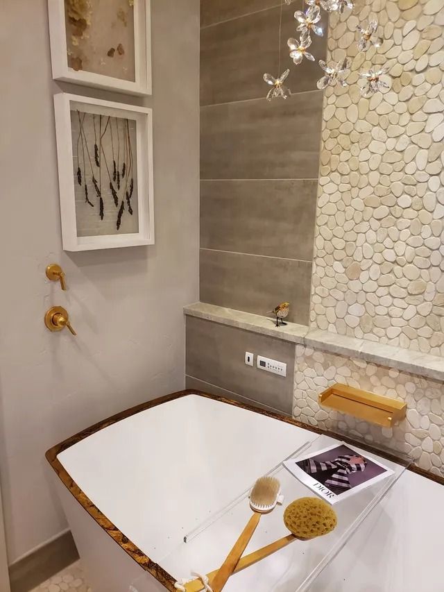 Bathroom with white soaking tub, pebble tile wall, artwork, and gold fixtures.
