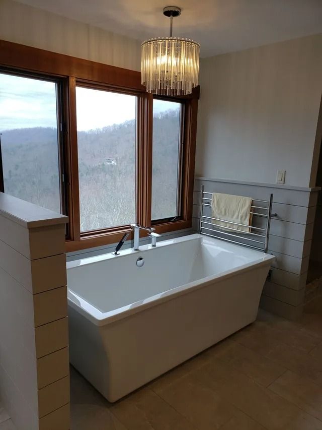 Bathroom with a white freestanding bathtub, large windows with a view, and a crystal chandelier.