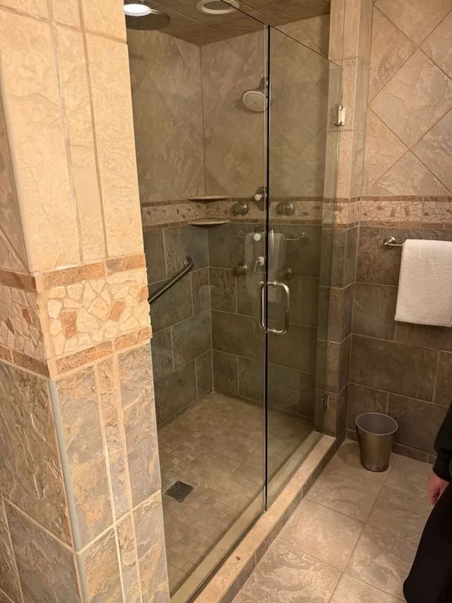 Shower stall with glass door, beige tile, grab bar, and recessed shelf.