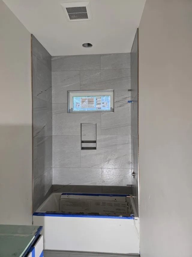 Bathroom with gray tiled shower wall, built-in shelf, and recessed window above a tub.