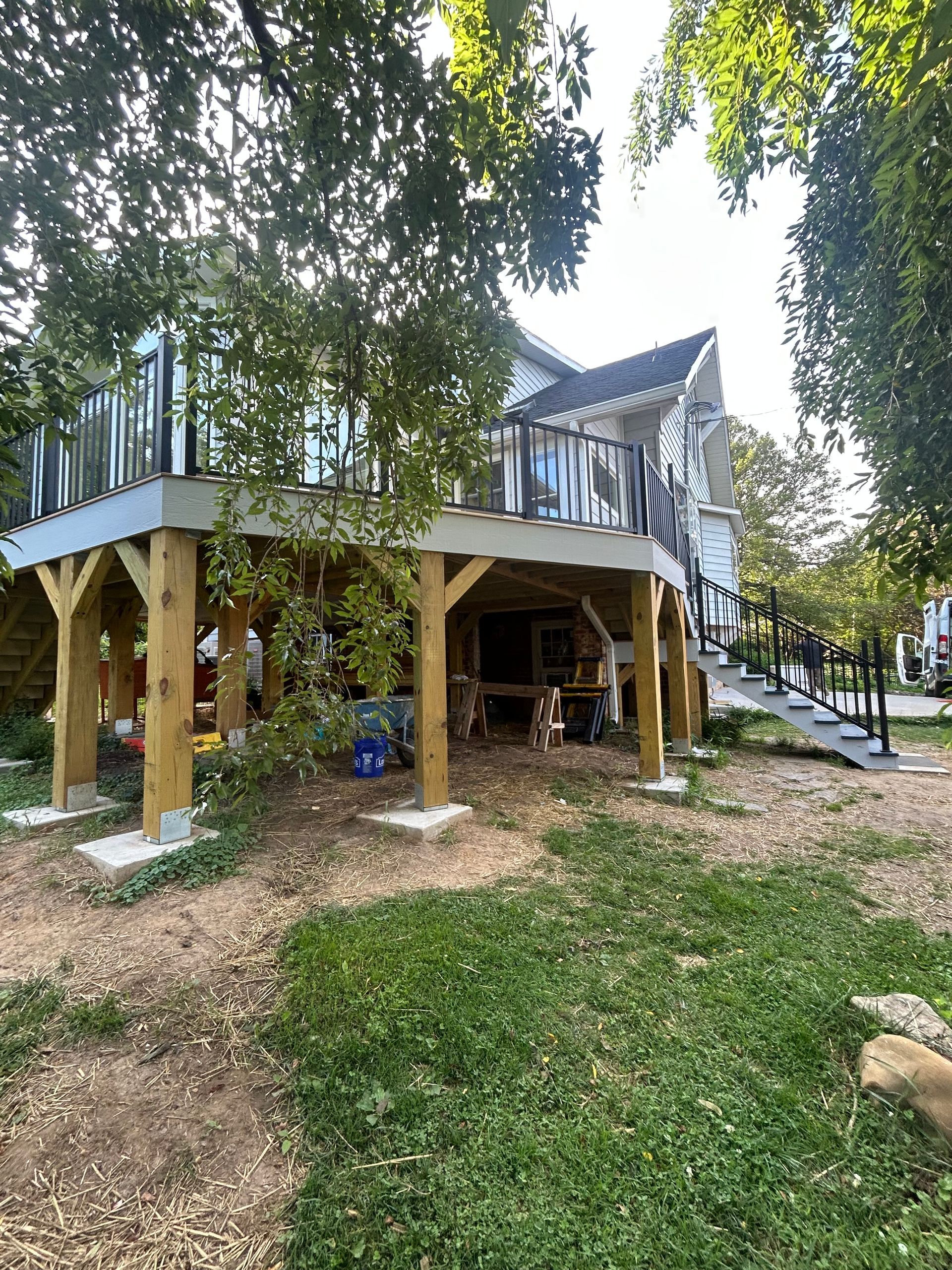 Stilted wooden house with open undercroft, surrounded by trees and grass, in a sunny yard