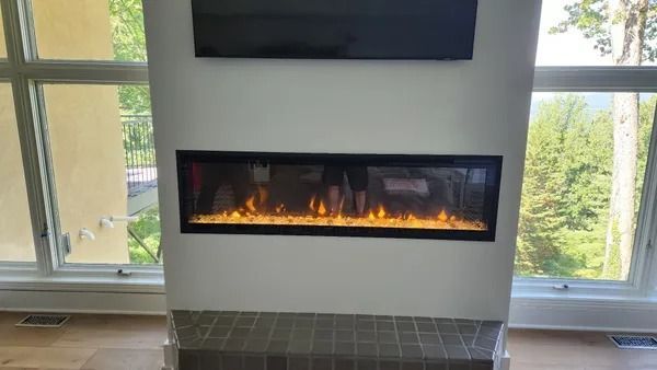 Electric fireplace with orange flames, set in a white wall below a mounted TV, next to large windows.