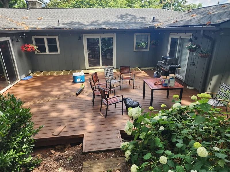 Backyard deck with furniture; gray house. Brown deck boards. Plants in foreground.