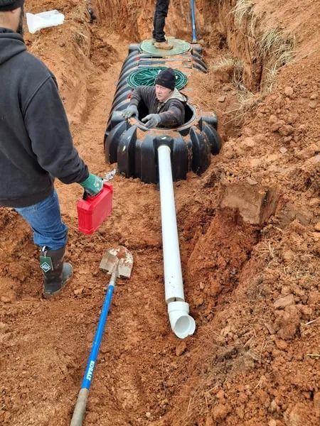 Workers installing a septic system. A man inside the tank, another holding a gas can.  Pipe and shovels in a dirt trench.