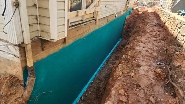 A trench next to a building wall with a teal-colored waterproof coating. Brown soil surrounds the trench.