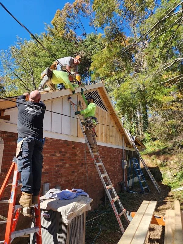 Construction workers installing siding on a brick building with ladders, sunny day, trees in background.