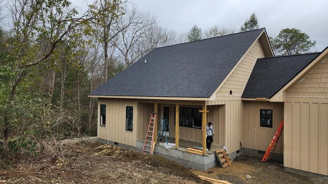 A house under construction with tan siding and a dark roof; a person stands near the porch.