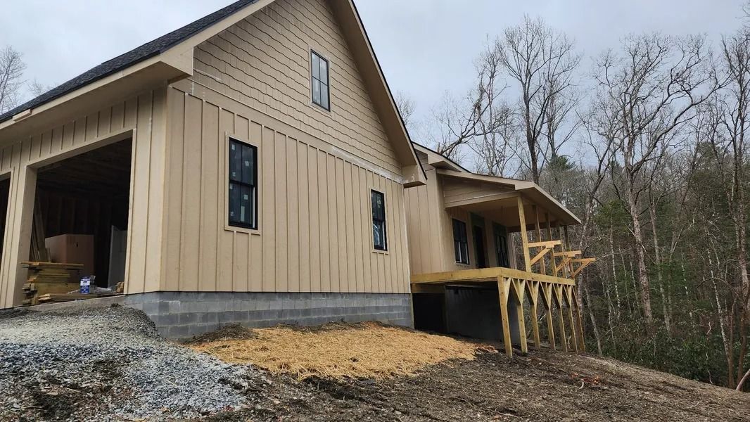 New house under construction with light-colored siding, a garage, and a porch, set in a wooded area.
