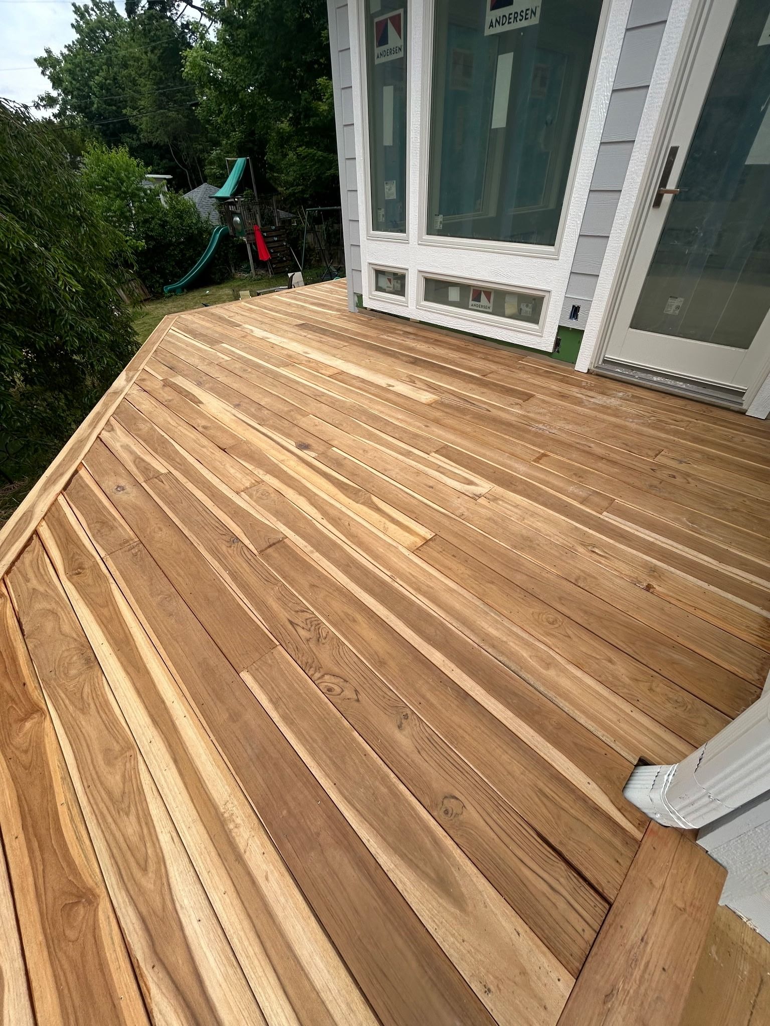 Wooden deck with white-framed glass doors beside lush green trees and a sloped railing.