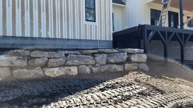 Stone retaining wall next to a light-colored house. Construction site with dirt and equipment visible.