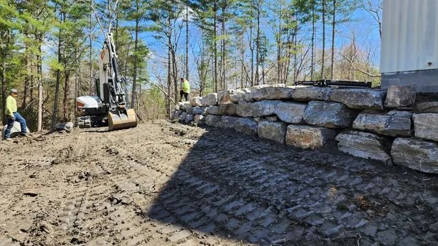 Construction site: excavator and workers building a stone retaining wall in a wooded area.