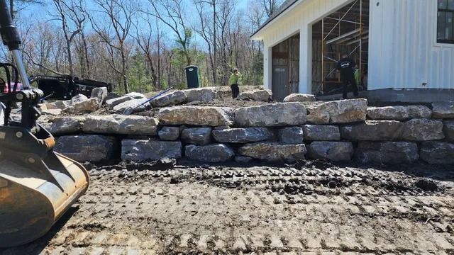 Construction site with stone retaining wall. Workers near a partially built white building with an excavator in the foreground.