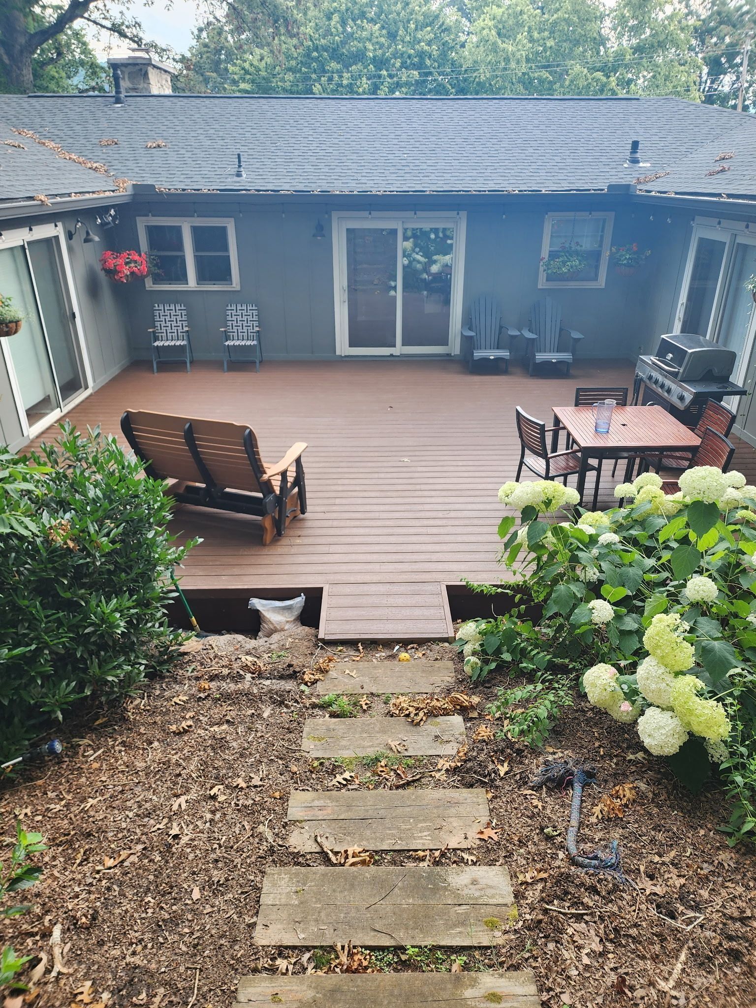 Backyard deck with furniture; stone path leads to it, surrounded by plants and a house.