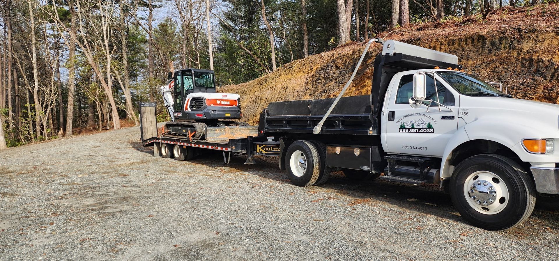 A white truck pulling a trailer with a Bobcat excavator on a gravel road, trees in background.